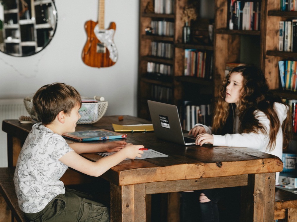 A brother and sister working together at a wooden table, books open and a laptop between them, in a warm room lined with bookshelves